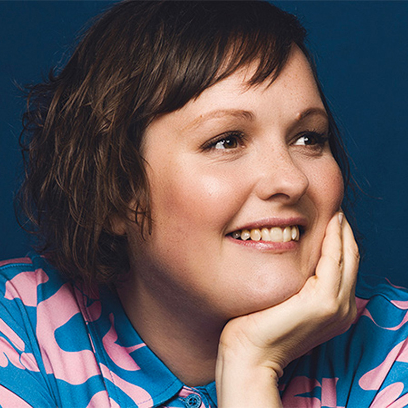 Josie is smiling looking to the right of the image with her chin in her hand, wearing a blue top with pink flowers on it. She has short brown hair.