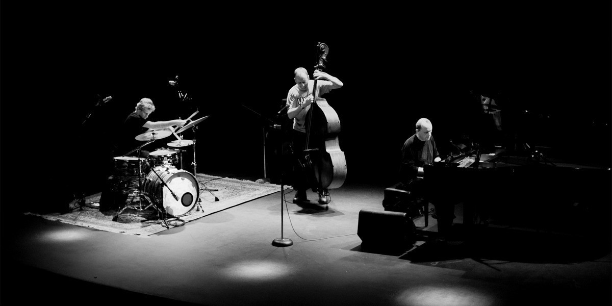 A drummer plays on a full drum kit on left hand side atop a rug, with a double bass player playing standing up in the centre and a pianist on a grand piano on the right hand side, all in black and white