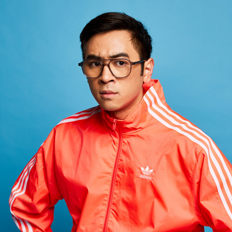 Michael Hing - Coming Out of My Cage and I'm Not Doing Well - Michael wears a bright orange tracksuit and glasses, he is sitting in front of a bright blue background. He has a pensive expression on his face.