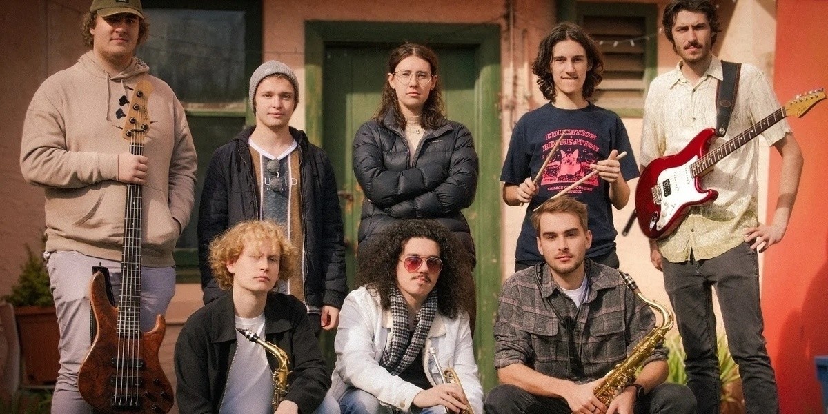 Eight band members stand together with their instruments in front of a green door.