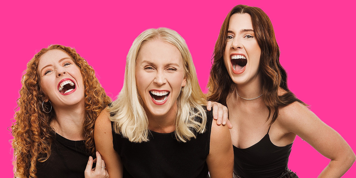 Three women, one with orange curly hair, one with blonde hair and one with brown hair, all dressed in black, laughing towards the camera on a pink background.