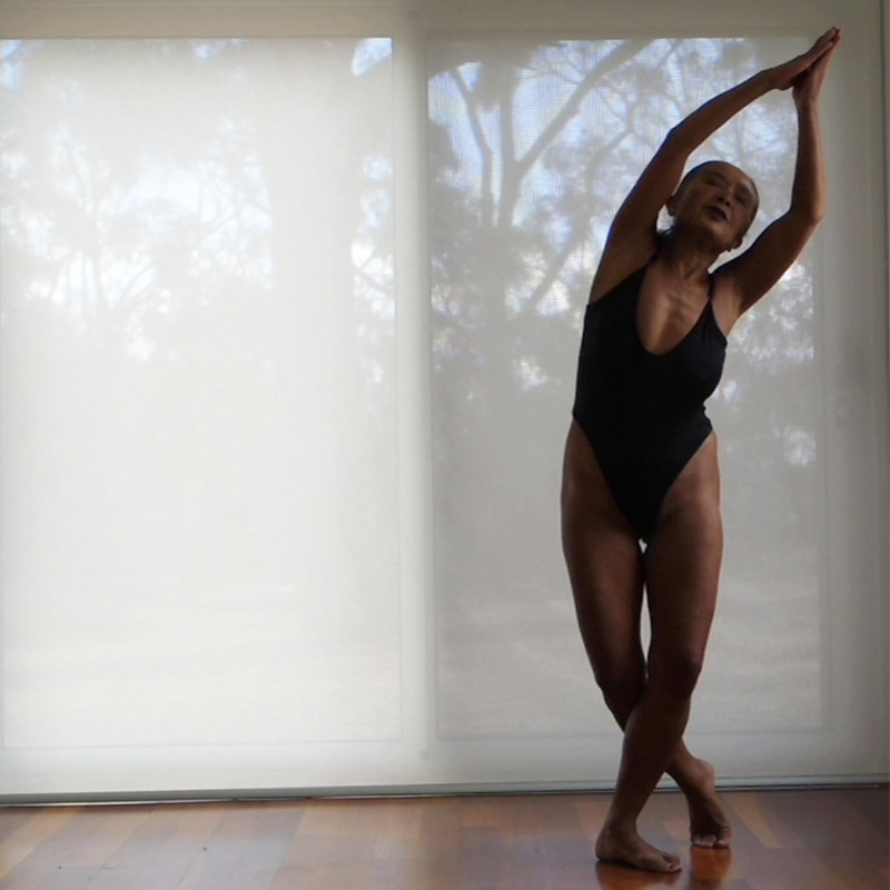 A photo of a person wearing a black leotard stretching. They have their arms above their head and one leg behind the other and are leaning to one side. The background features a wooden floor and sheer white curtain covering a window.