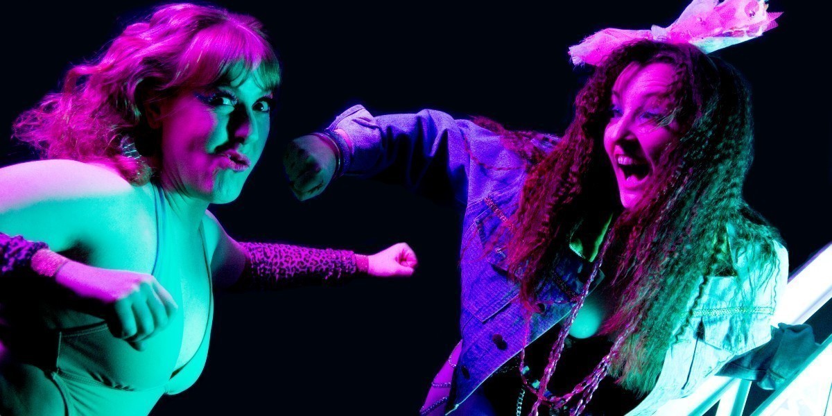 Two burlesque wrestlers in 80’s style outfits are lit by green and pink lighting against a black background. One performer is standing on a ladder and smiling gleefully while throwing a haymaker punch to the other’s jaw. The other performer is looking at the camera while pulling a silly face from the implied impact of the punch and has their arms out to either side.