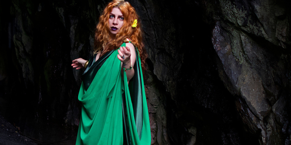 A white women with red curly hair wears a draped green gown stand with her body turned to the right while reaching her left arm out to the viewer and making eye contact.