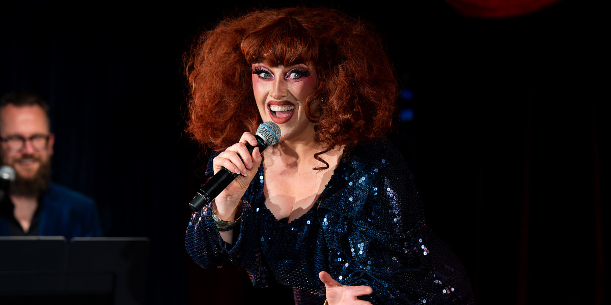 Glamorous red-headed woman in a short blue sequinned dress smiles while holding a microphone, her male accompanist laughing in the background while sitting at an electric piano. Photograph by Keiran McNamara.