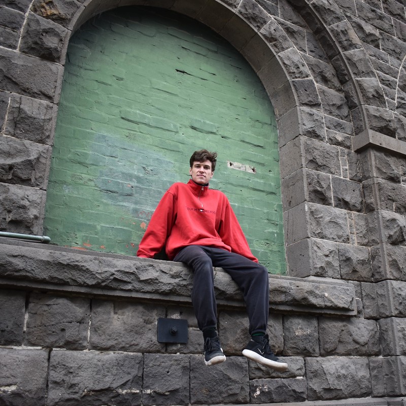 A young man sits on the ledge of an archway on a large grey stone wall. The arch is bricked up and painted green. He wears a red high necked jumper, black tracksuit pants and sneakers