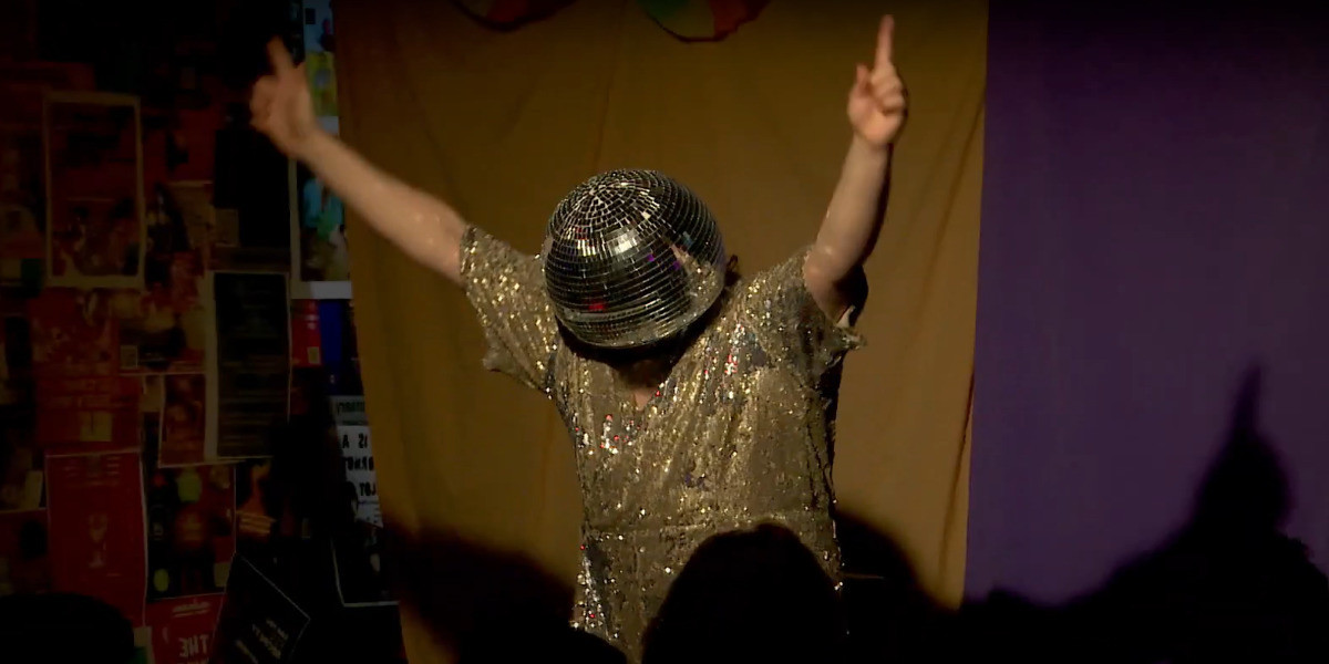 Mr Disco has their arms raised - they are wearing a disco ball helmet and a silver sequined jumpsuit