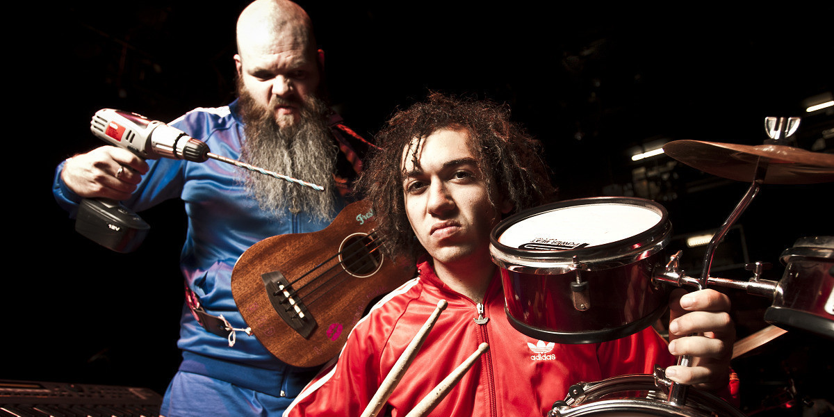 Adam Page wearing a blue tracksuit holds a ukulele in his left hand and a drill in his right hand pointed menacingly at Gene Peterson who holds drumsticks in his right hand and a tiny drumkit in his left. Both artists are sneering