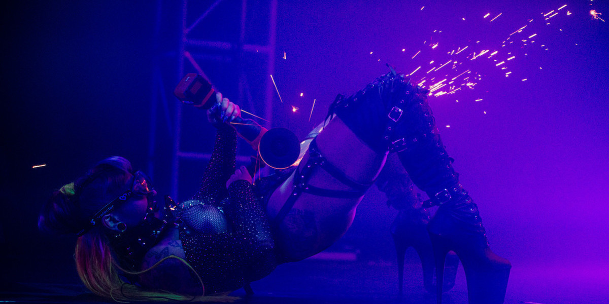Neo-punk style side-show performer with neon hair and safety googles is lying on their back on the stage, holding an angle grinder to a metal plate attached to their costume. Bright sparks are flying from the grinder across the stage, contrasting against dim purple and blue stage lights.