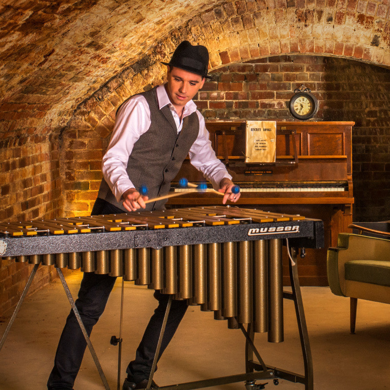 A photo of a man playing a vibraphone. The man is wearing a black hat, pink long sleeved shirt and brown vest. The background features an old red brick wall and ceiling and a wooden piano sitting in the background.
