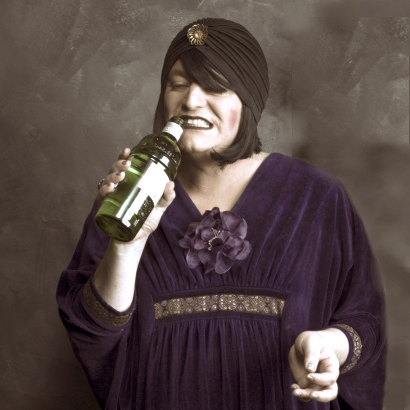 Female presenting 1920s style singer with a gin bottle as a microphone, dark purple dress on and dark rouge and lipstick. She is wearing a turban over her black bob haircut.