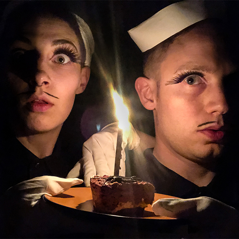 Two boys are holding an orange plate with one slice of birthday cake on it.  Both boys are wearing white sailor hats, one large fake eyelash, and have thin pencil mustaches.  The piece of cake has one lit birthday candle stuck into it.