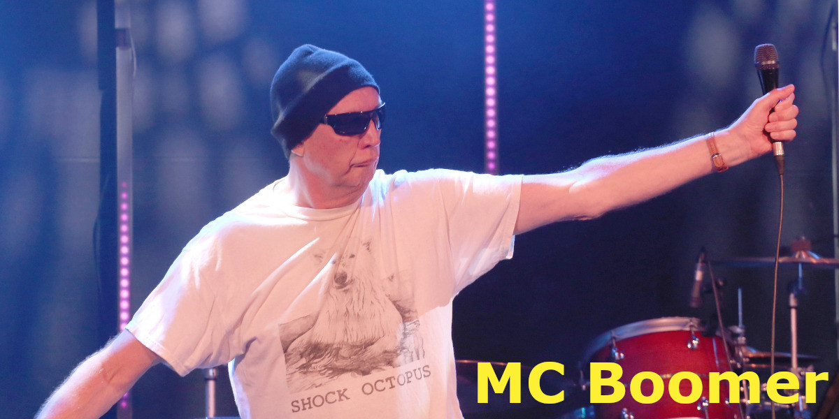 MC Boomer holding a microphone on his left hand. He wears white t-shirt, black beanie and black sunglasses.