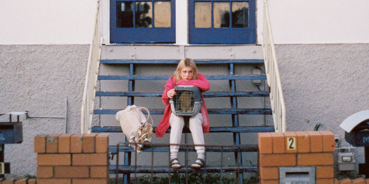 A young woman with bleach blonde hair sits on the steps of a house. She has a cat carrier on her lap, containing a ginger cat. She is wearing a pink hoodie, tracksuit pants, and Birkenstocks with socks. Next to her on the steps is a duffle bag, with sparkly gold stripper heels strapped to the handle.