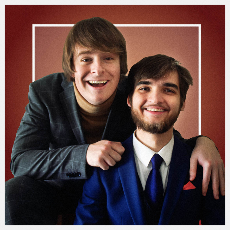 Aiden Willcox and Isaac Haigh wearing 70s era suits and grinning cheesily toward camera on a maroon background.