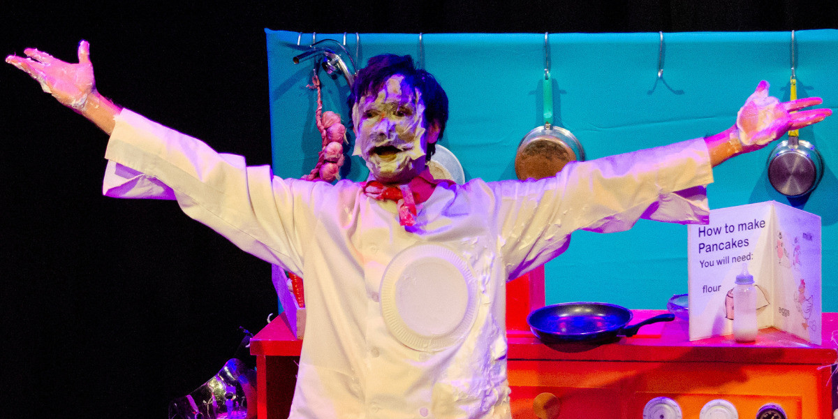 A man is standing with his arms spread wide. His face is covered in cream from a cream pie. A cream pie is also attached to his shirt.