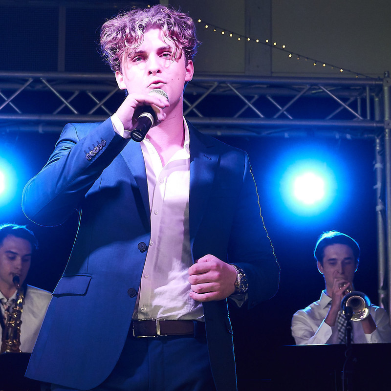 A photo of a man singing into a microphone on stage. He is wearing a blue suit and white shirt. He has blonde tousled hair. In the background behind the singer, a saxophone and trumpet player are performing.