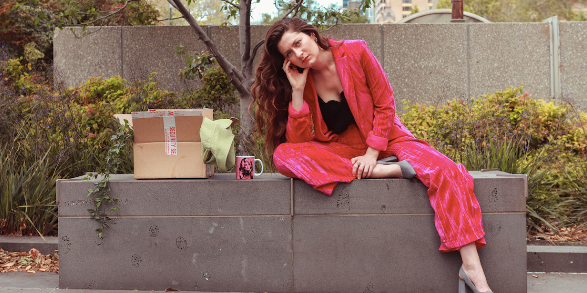 Image of young woman wearing a pink velvet suit, sitting on a concrete bench, next to a box containing a green jacket and wine bottle.