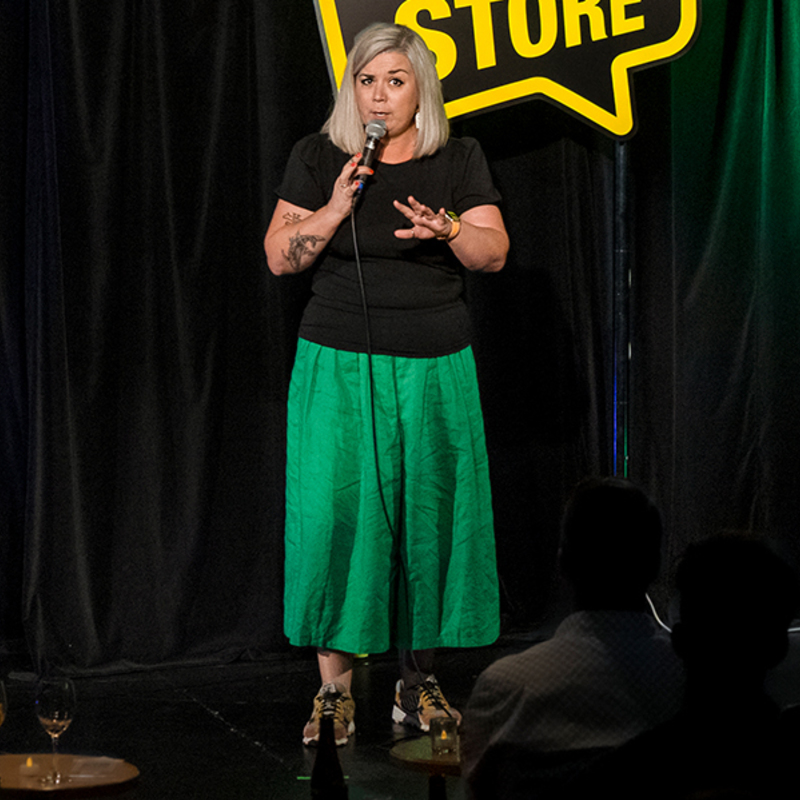 Image of Comedian, Nicky Wilkinson, live on stage, at the Comedy Store in Sydney. She is wearing a black top, green baggy trousers and colourful trainers. She is holding a microphone.