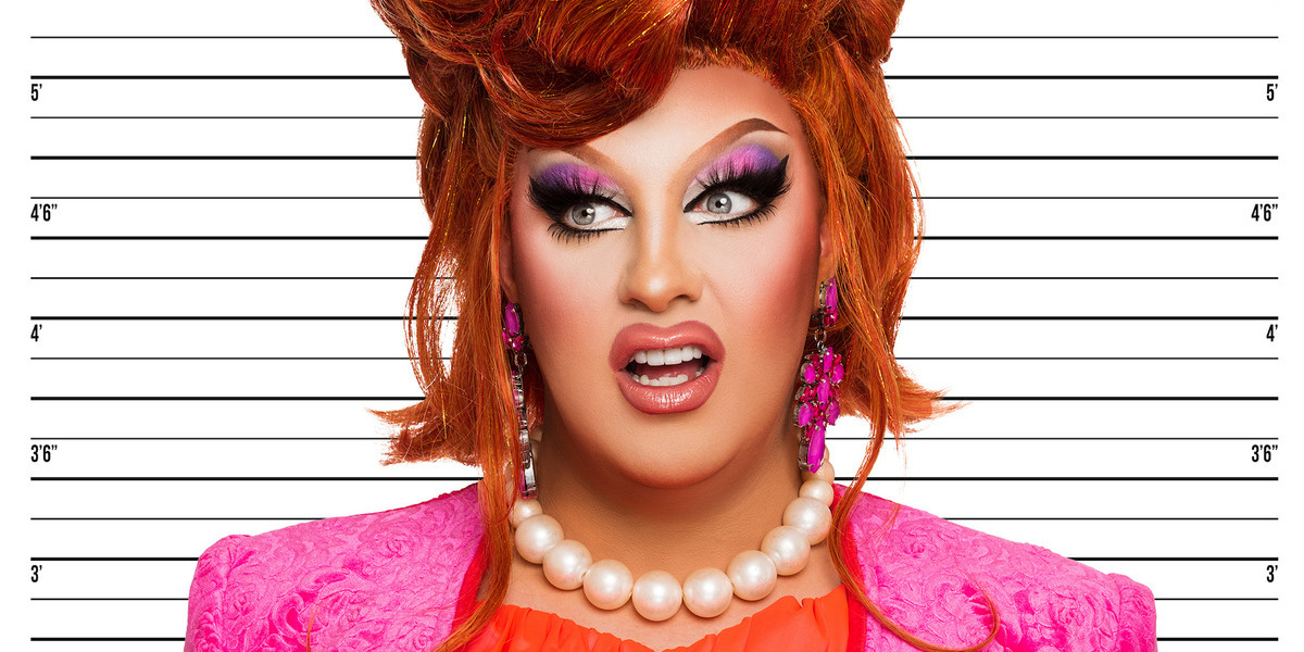 The Artist, Karen From Finance, stands in front of a police photo wall while she gets her "mug shot" portrait taken. She stands at seven foot tall with her curled, auburn, beehive hairstyle. She is wearing a pink power suit with an orange under shirt, a large pearl necklace, and pink pendant earrings. She is standing front on and staring slightly to the right of the camera with a shocked expression on her face.