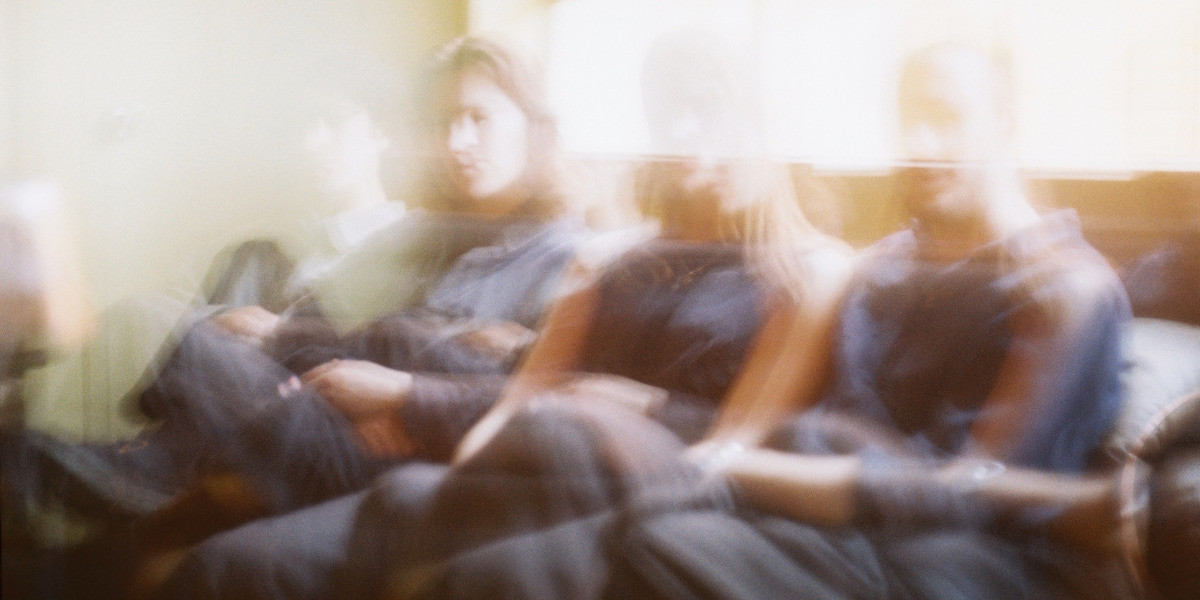 Blurry image of band members sitting side by side on a couch, soft light coming through the window.