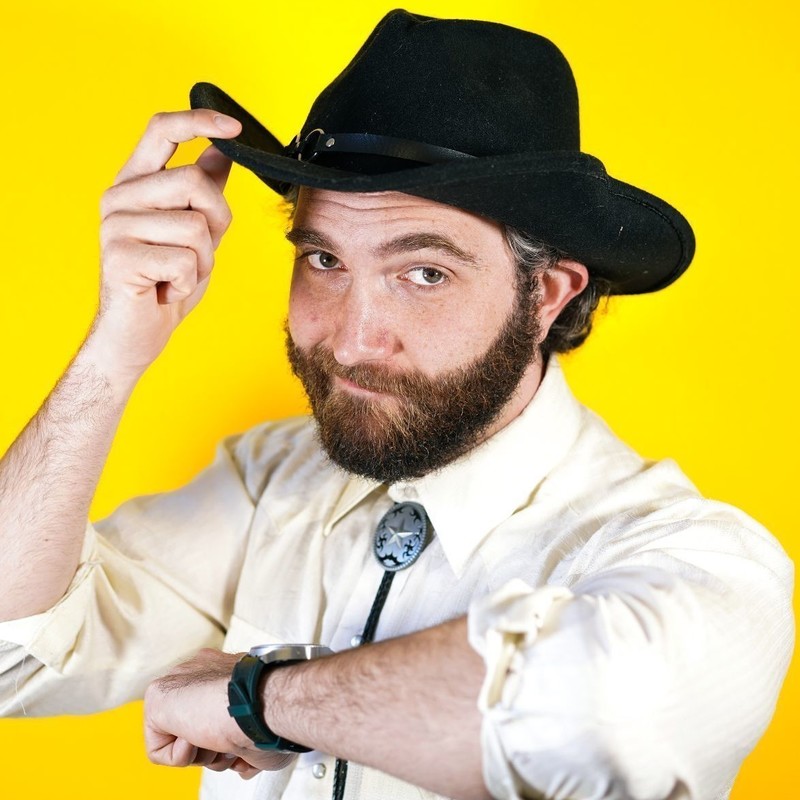 Comedian Jacob Jackman. He is a white CIS man with a beard. He is wearing a bollo tie and a cowboy hat, tilting it slightly with one hand. His other arm is angled so it looks like he is leaning on the frame of the image.