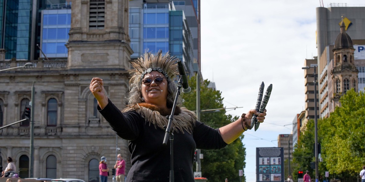 Kaurna Elder, Katrina Power, holds up tapping sticks and wears a headress. She is addressing the crowd.