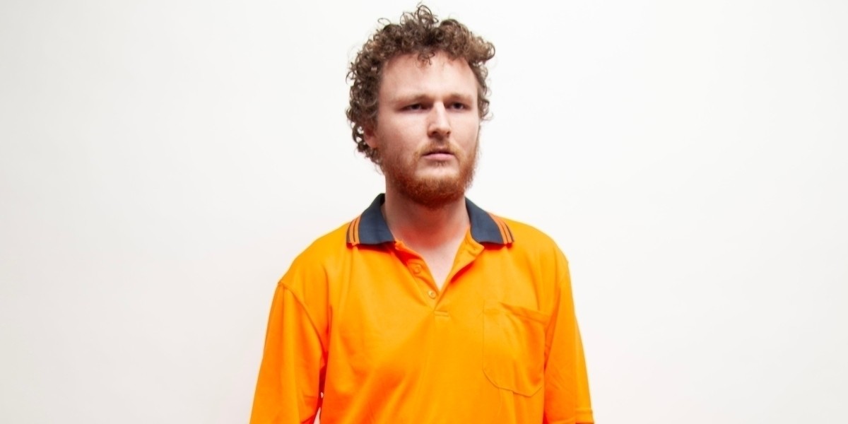 Comedian Jack Knight staring ahead off camera with a blank expression, wearing an orange hi-vis collared shirt.