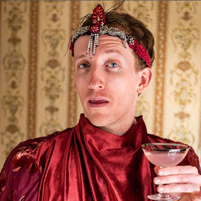 Table for Two performer, Will Tredinnick, faces the viewer dressed elaborately as a Maitre de. Draped in red silk, and holding a pink cocktail, he wistfully looks over his shoulder.