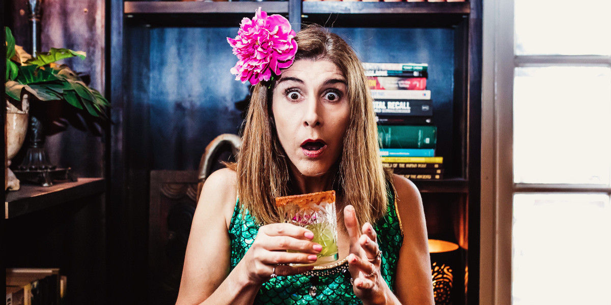 A woman with shoulder length brown hair, with a pink flower in it, is has a cocktail glass held up just below her mouth. Her lips are parted in a shocked expression, and her eyes are wide. Behind her is a bookcase.
