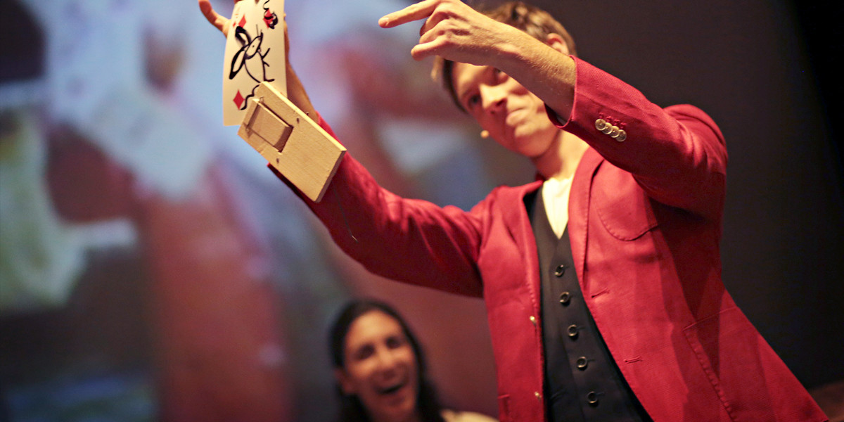 Magician Martin Brock in a red jacked, holding up a rat trap with a spectators selected card. The spectator is in the background with an amused facial expression.