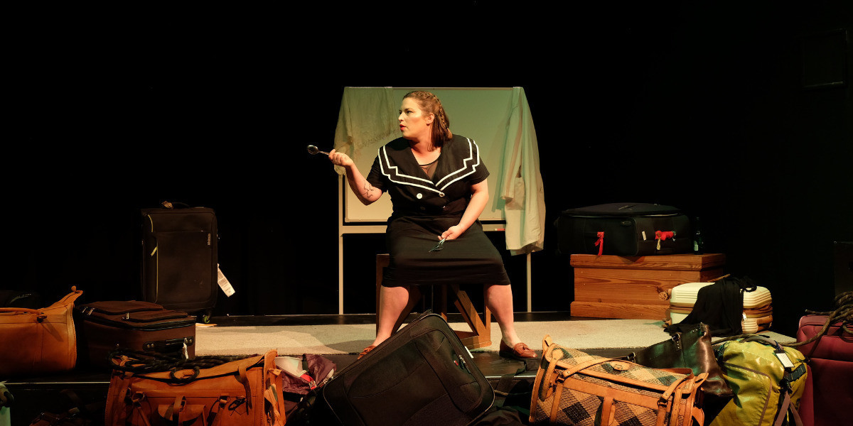 Wide shot. Woman sits amongst bags on stage, in a 50's style dress. She is holding a fork and knife in one hand and pointing with a spoon in the other, to her right.