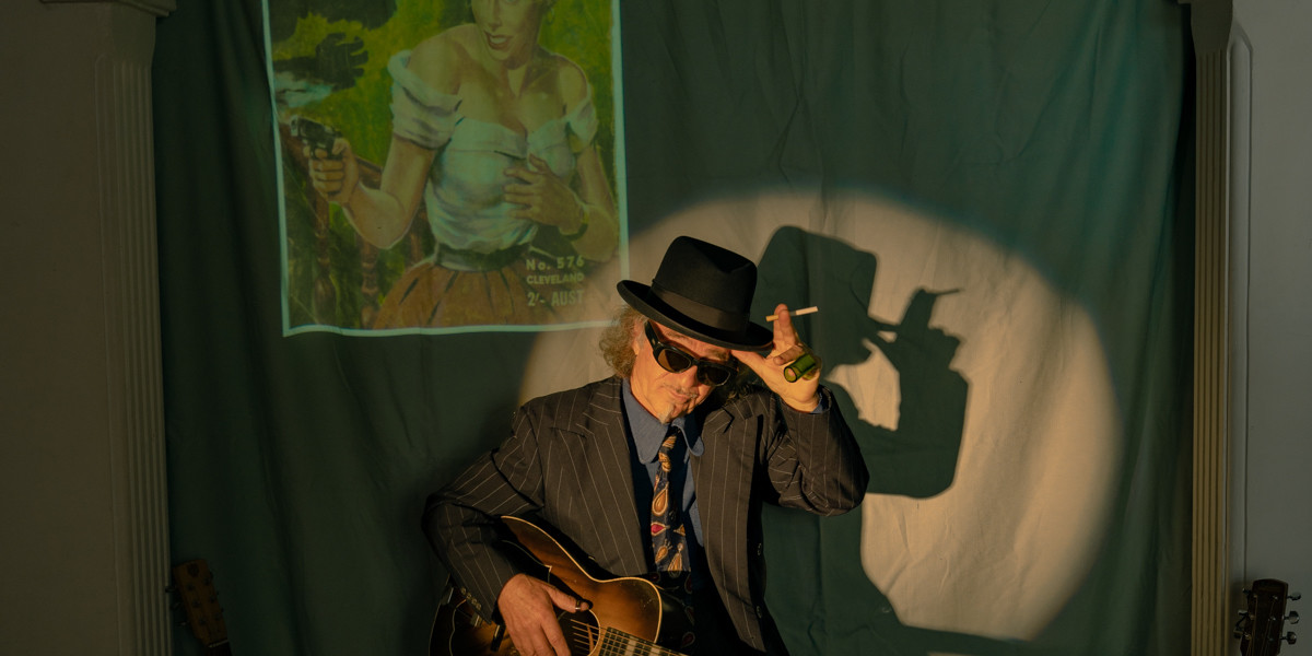 a man wearing a suit and  sunglasses is tilting his hat with a hand holding an unlit cigarette. In the back ground there is a poster picture of a woman shooting a gun at the shadow of a man