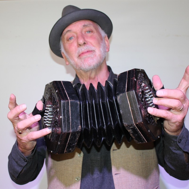 A photo of a man wearing a black hat, grey shirt and cream coloured vest holding a hand held accordion.