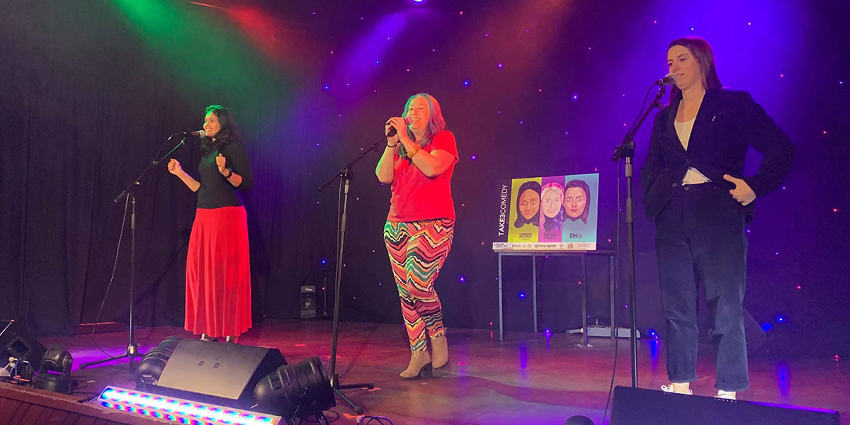 The performers of Take 3 on stage. Bathed in blue, red and green lights against a starry background, they stand equidistant apart in front of boom mics. Behind them is the Take 3 poster, featuring illustrated versions of each performer.