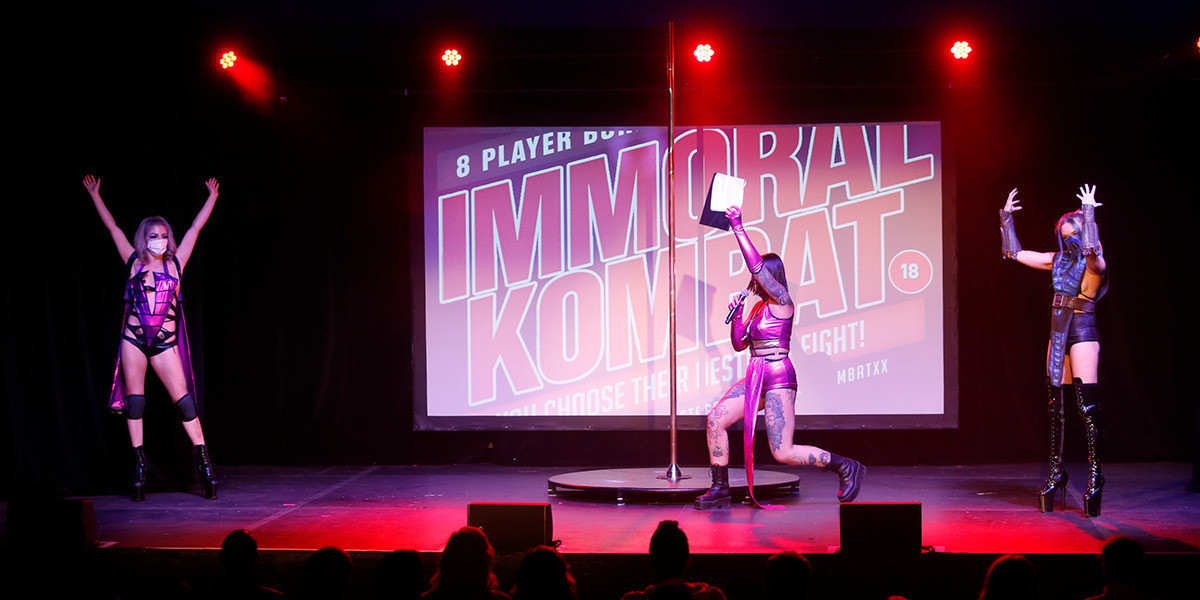 Saskia as Mileena waves a clipboard between Fable Fireside dressed as Sindel on left and Vauxx as Sub Zero on the right of a stage. A pole dancers pole in the centre of stage.