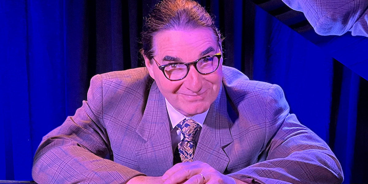 Doctor H, a middle-aged causacian male wearing glasses and wearing a fine check suit, is leaning over his piano, smiling at us with his eyebrows raised