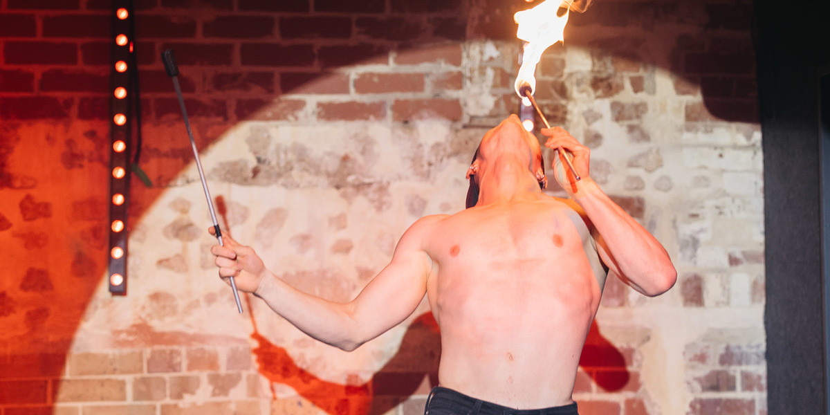 Super hot performer with no shirt facing the sky whilst breathing out fire. The backdrop is stone and brick wall with nightclub lighting and the shape of a pin spot light.
