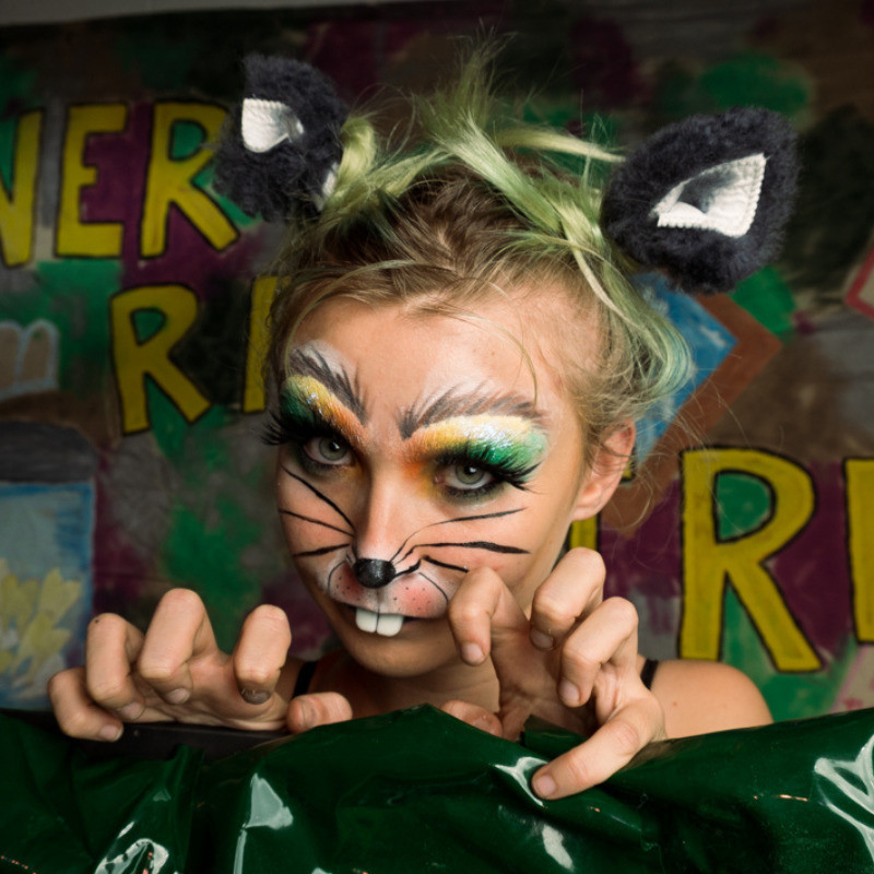 Sewer Rat Girl - Picture of Sewer Rat Girl (wearing rat ears and buck teeth). She is peering over a green flat with her hands. Sewer Rat Girl is written in yellow on the backdrop behind.