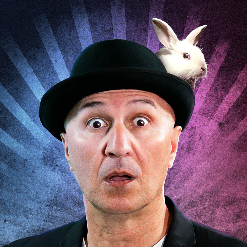 TAHIR - THE WORLD'S BEST WORST MAGICIAN! - An image of a man with a shocked facial expression. He is wearing a black hat and there is a white rabbit poking its head out the side.