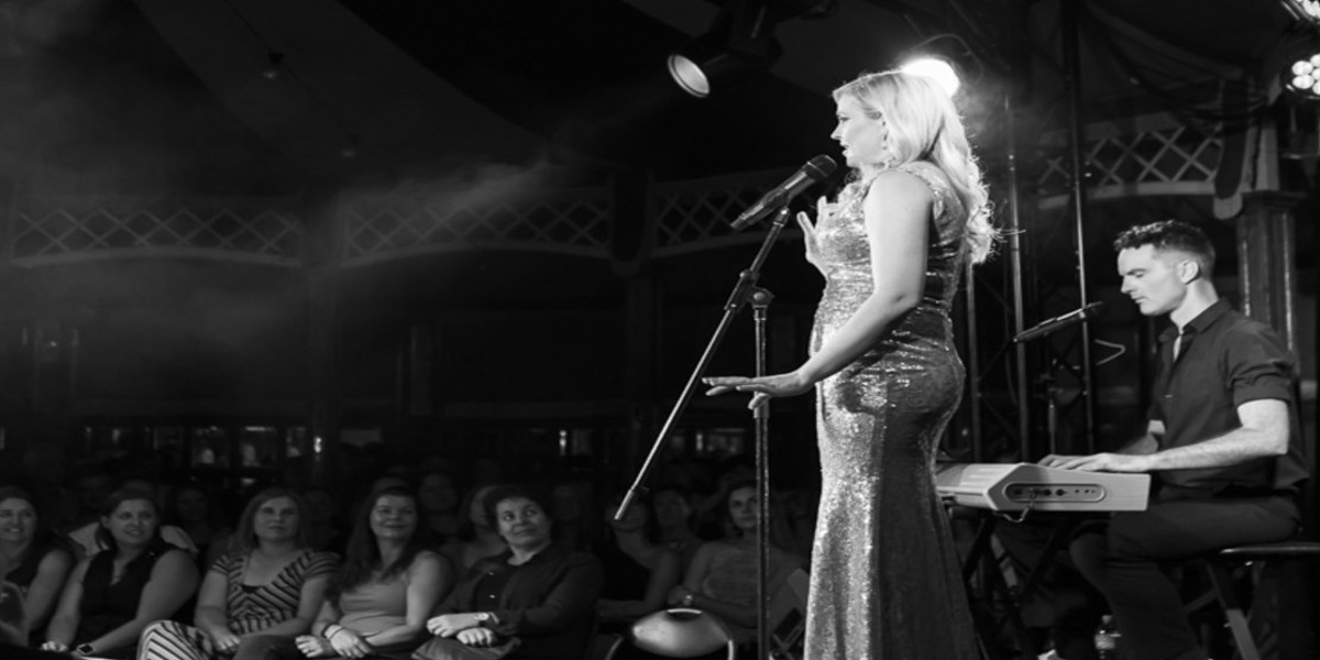 A black and white photograph of a woman with medium length blonde curly performing on stage. She is wearing a sparkly silver dress and singing into a microphone. In the background there is a seated audience, and a man seated at a keyboard behind her.