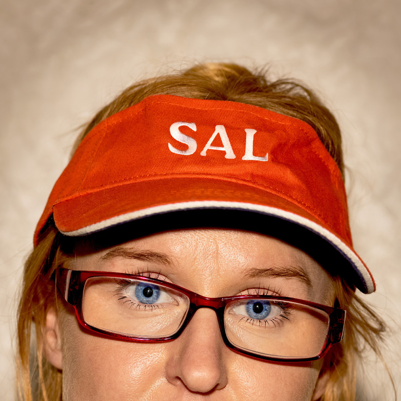 A close up of a woman's face from the nose up wearing red reading glasses and orange visor with the name Sal written on it in white. She is looking forward directly at the camera with a white textured wall in the background. Her expression is direct and slightly scathing.