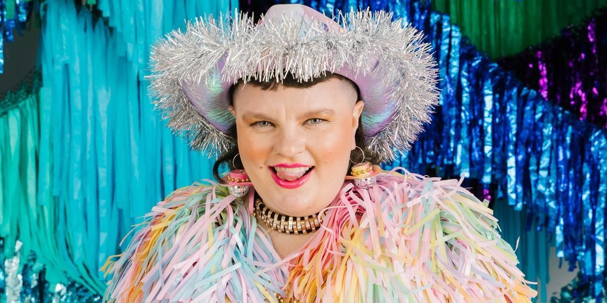 Annie Schofield is posing in front of a backdrop of blue and green shimmery streamers. They are wearing a silver Cowgirl hat and a pastel pink tinsel jacket. They smiling mischievously at the camera with their tongue sticking out a little bit