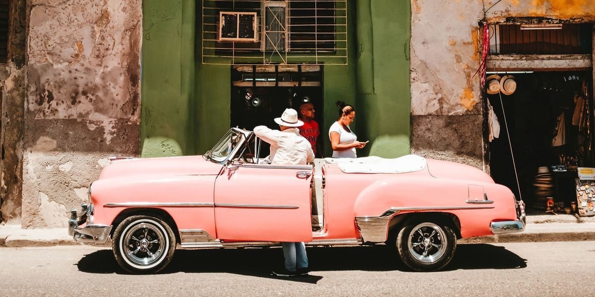 Man with a hat standing by his pink Cadillac car looks into a Cuban shop. A women on the footpath is texting. Another man walks out of shop in a hurry.