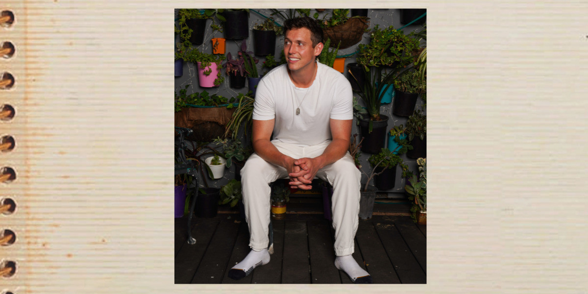 Colin sits on a stool dressed in all white in front of a wall of plants as he looks off to the side smiling