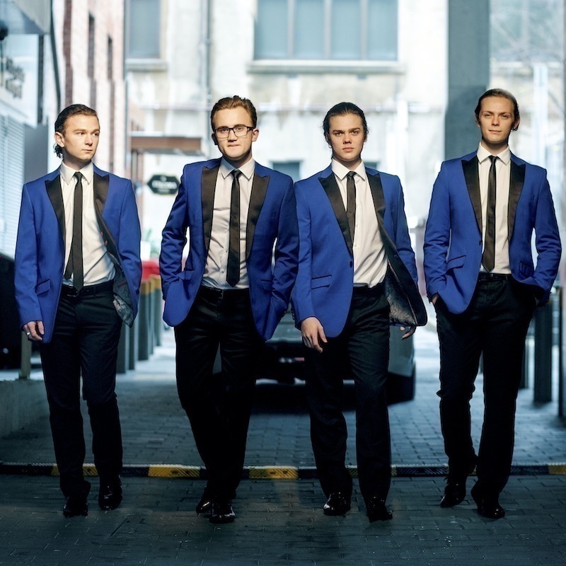 A photo of four men walking with blank expressions on their faces. They are wearing matching blue suit jackets, black pants, black ties and white shirts.