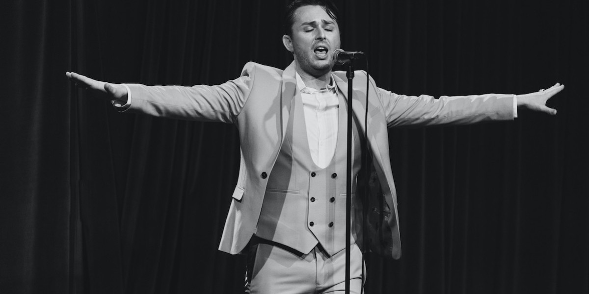 Black and white image of a man singing passionately into a microphone holding his arms outstretched.