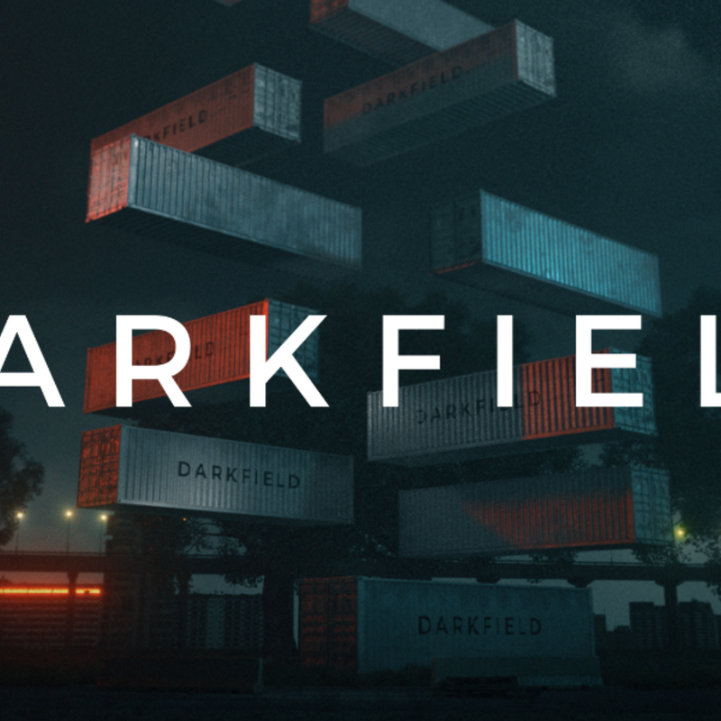 A dark landscape with shipping containers floating up in the air. It looks as if there was a stack of nine of them and they have begun floating upwards, leaving only one still on the ground. Each container has the word DARKFIELD written on it. There are some streetlights and trees on the horizon in the background.