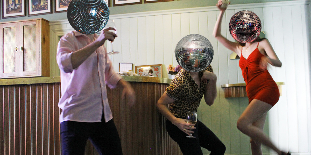 Three people dance in front of a bar. They wear a long sleeve pink shirt, an animal print shirt, and a red dress. They have disco balls instead of heads. They dance as individuals, not touching. Their movements appear slightly blurry as they twist into various shapes, creating angles with their arms and legs.