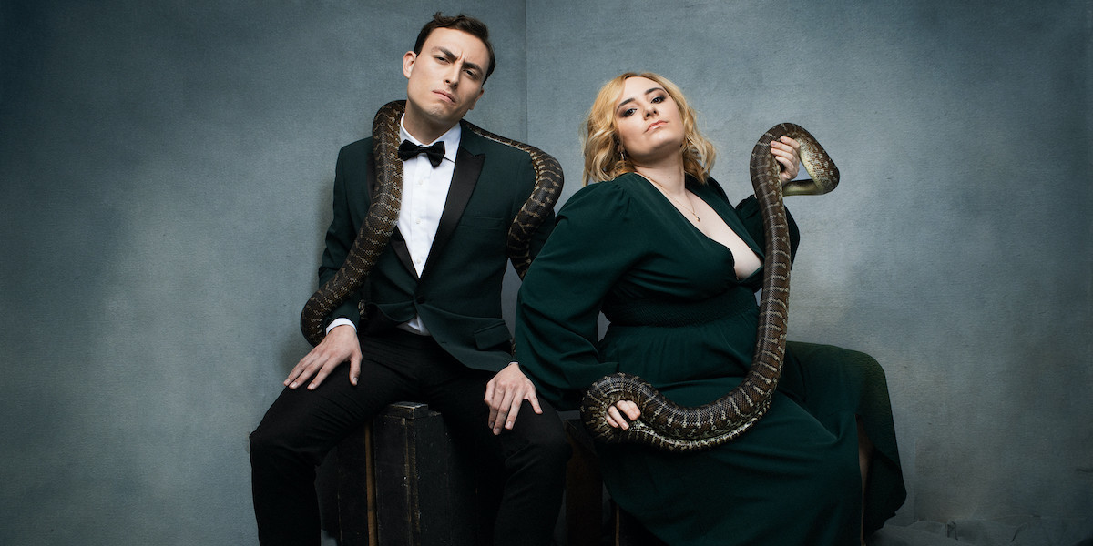 The cast of Snake Pit is posed in front of a grey background. Maddie Houlbrook-Walk and John Robles sit next to each other in green formal wear with snakes draped on their shoulders. They have serious smug expressions on their faces.